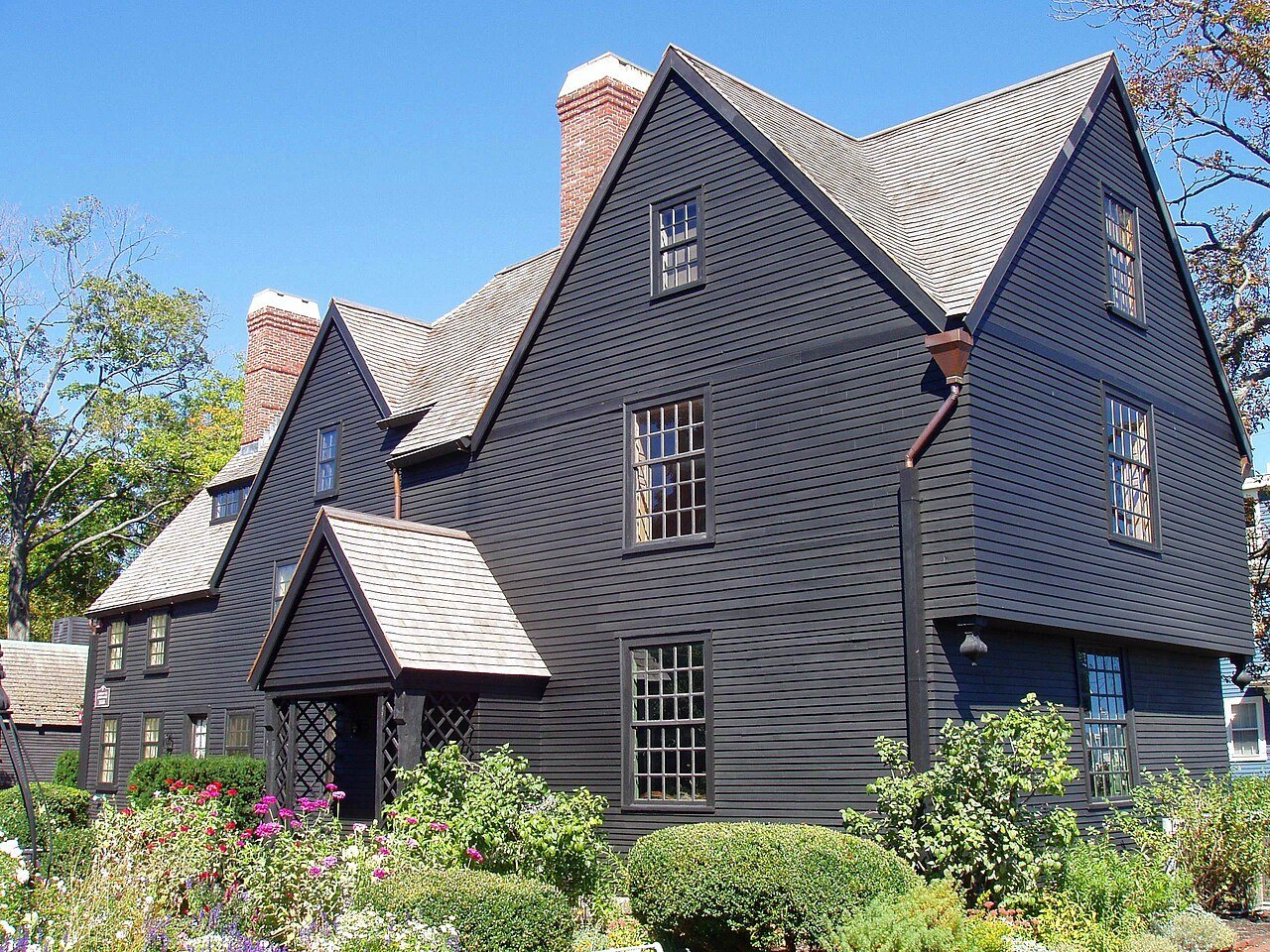 The dark clapboard House of the Seven Gables in Salem, with steep pointed gables, brick chimneys and a flower garden in front under a blue sky