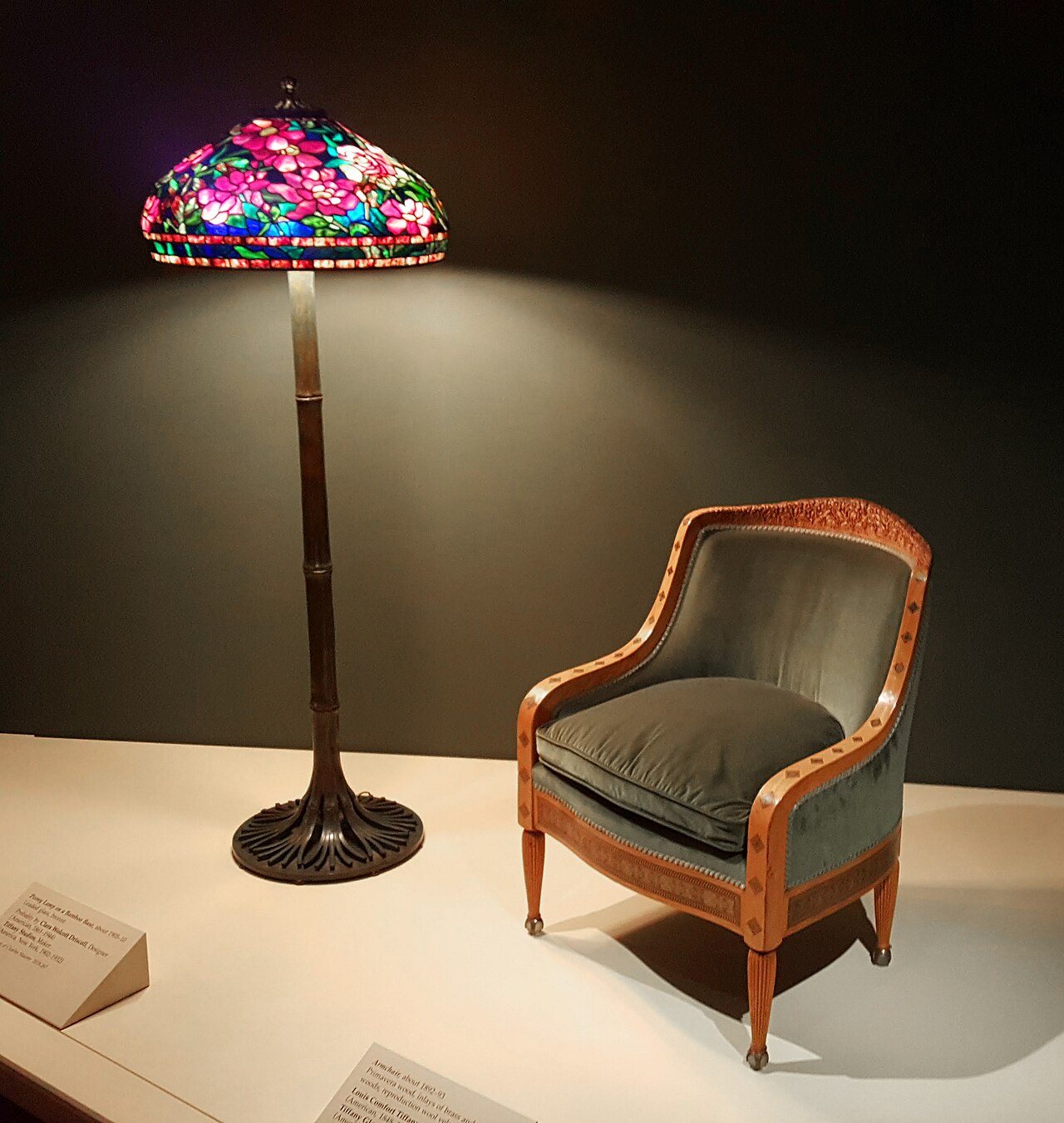A Tiffany Studios floor lamp with a glowing peony glass shade beside an upholstered armchair, displayed in a dim museum gallery
